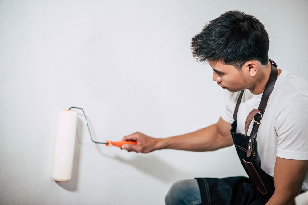 A person in an apron uses a roller to paint a white wall.