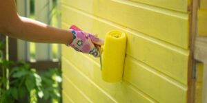 Person using a scraper tool to remove old, peeling yellow paint from an exterior wall surface