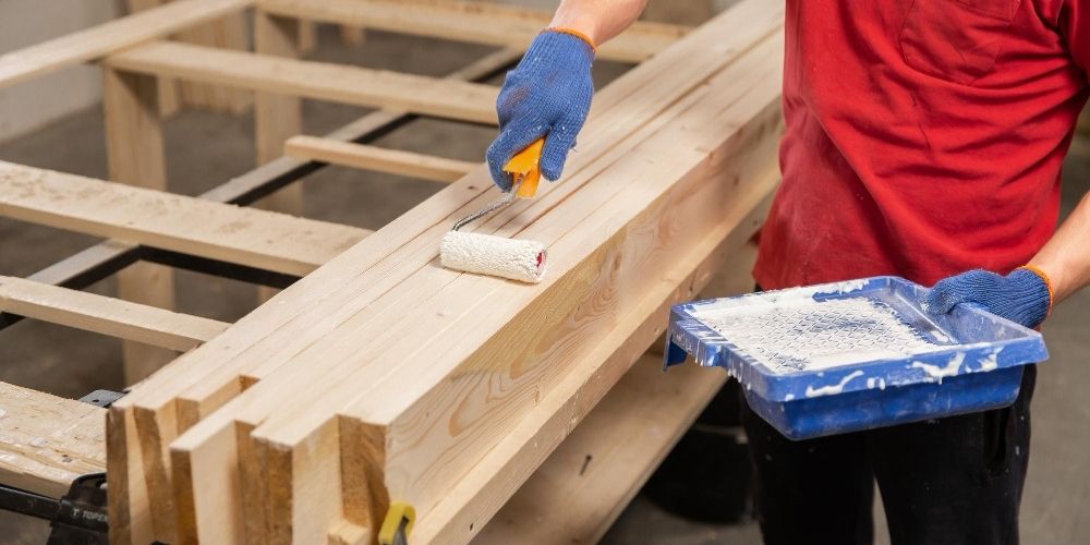 A person using a paint roller to apply primer or paint to wood siding on the exterior of a building